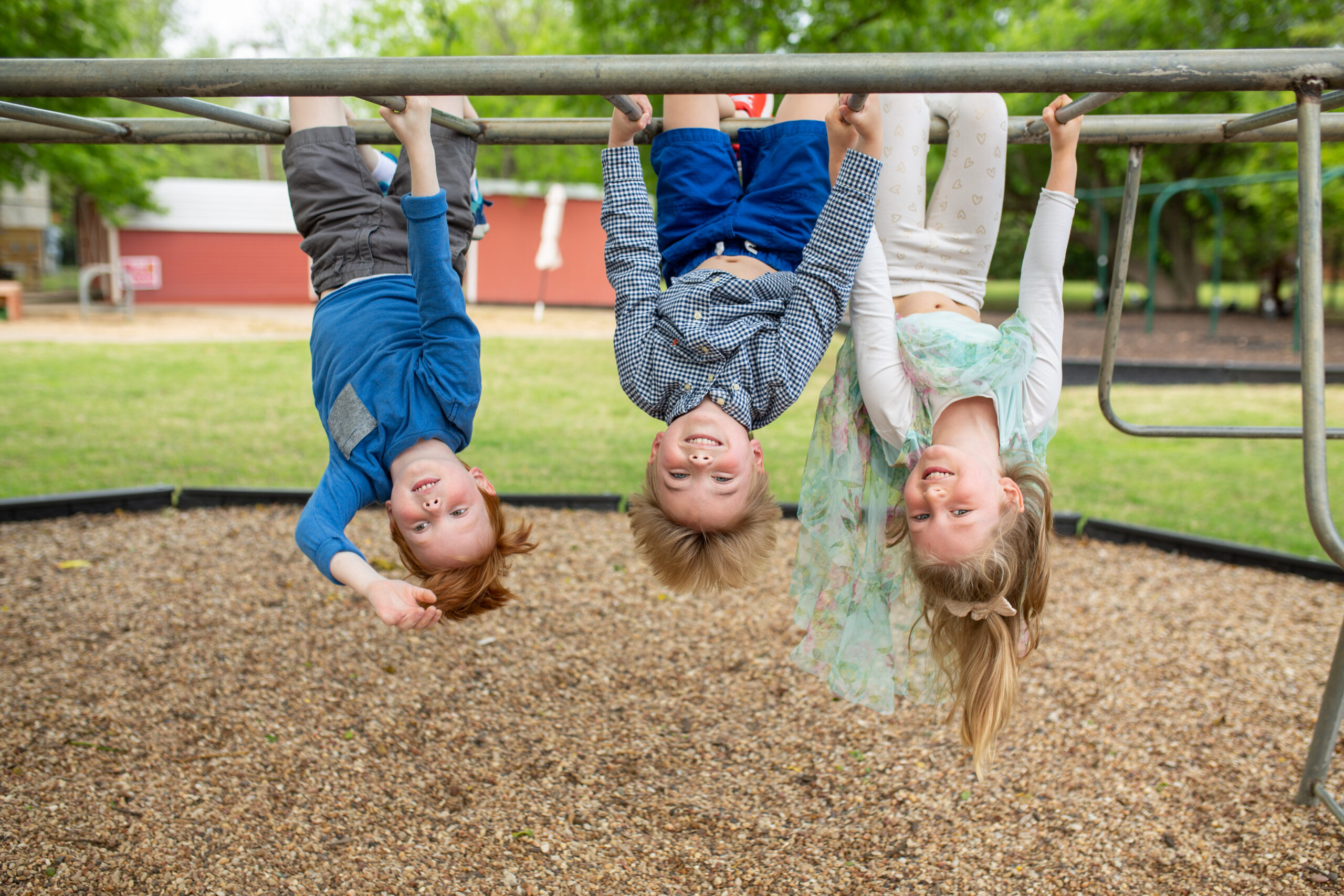 Three children hanging upside down from a bar on the playground at Northaven Co-operative Preschool in Dallas