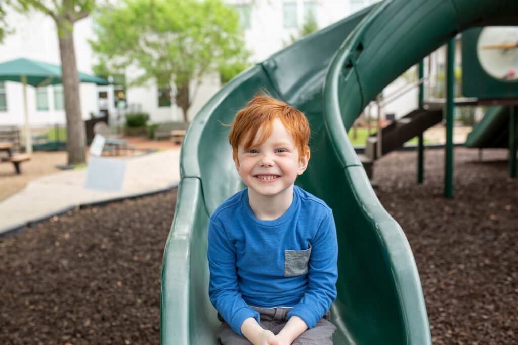 A boy with a blue long sleeve shirt is sitting on a green slide on the playground at Northaven Co-operative Preschool in Dallas