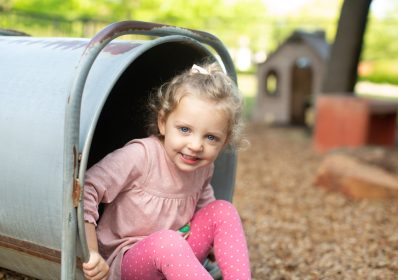 A girl wearing pink clothes is sitting at the edge of a crawl tunnel on the playground at Northaven Co-operative Preschool in Dallas