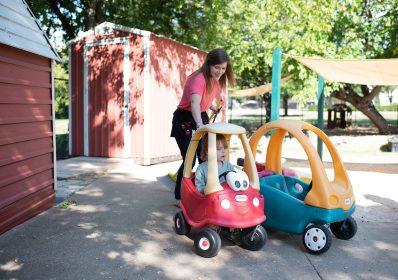 A teacher is guiding a student in a Little Tikes car on the playground at Northaven Co-operative Preschool